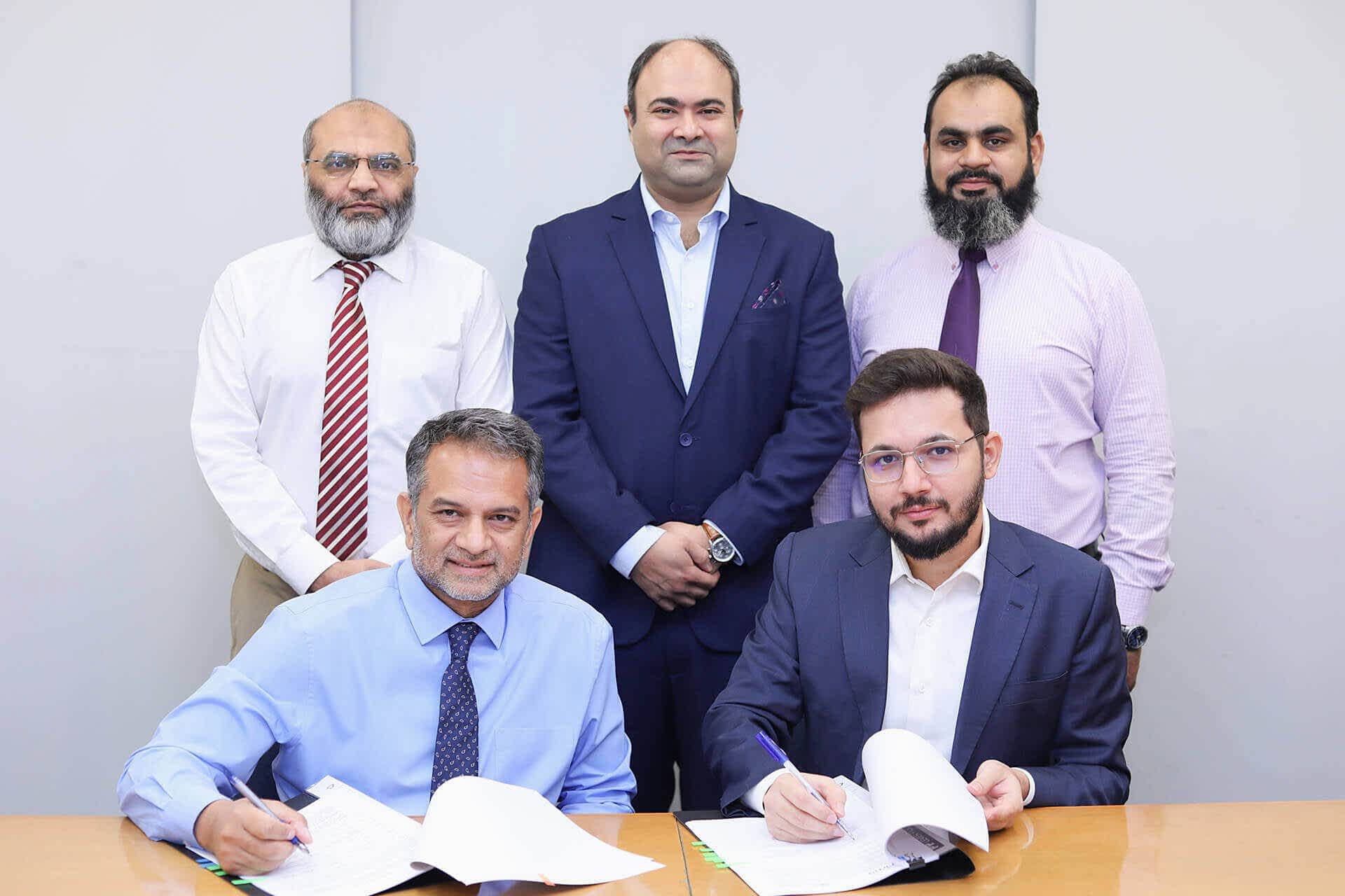 The image shows a formal signing event between Atlas Insurance Limited and Prima Consulting. The individuals in the foreground are Sumair Sayani (Prima Consulting’s Director of AI and Tech) and Baber Mirza (Atlas Insurance’s Chief Executive Officer), who are signing documents. Behind them stand three more key figures: Ibrahim Ahmed Zahidie (Director of Financial and Accounting Advisory), Omer Yousuf (Assistant Vice President), and Baber Mirza (again, the CEO of Atlas Insurance).