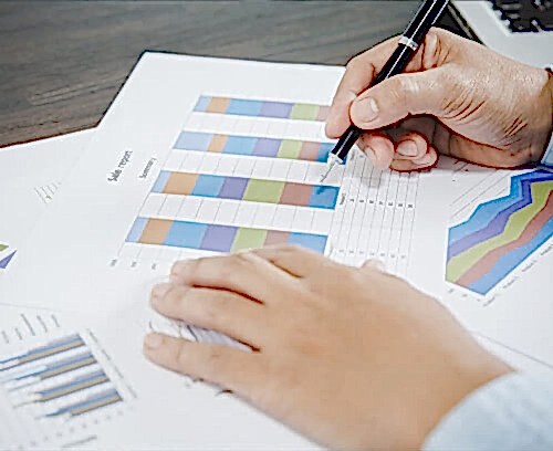 An individual’s hand holds a pen, pointing to a colorful bar graph on a printed paper. Additional graphs and data analysis charts are visible on the desk, conveying a professional setting where actuarial services involving data interpretation and financial analysis are being conducted.