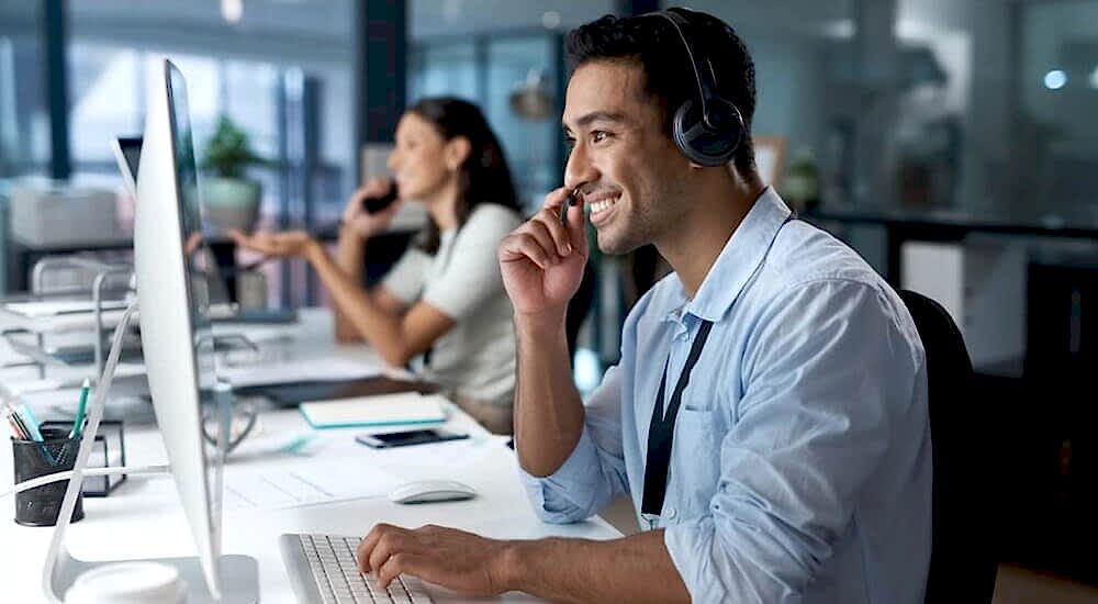 Two professionals in an office setting: A customer support representative wearing a headset interacts with a computer screen in the foreground, while another individual works on their computer in the background. The image conveys a professional environment where dedicated support is provided, aligning with the accompanying text about comprehensive assistance from actuaries and accountants through IAS 19 software