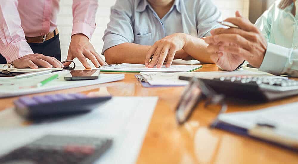 A group of professionals is engaged in a financial analysis, working with documents and a calculator on a table. The focus is on their hands and the papers, indicating a collaborative effort in reviewing detailed financial data.