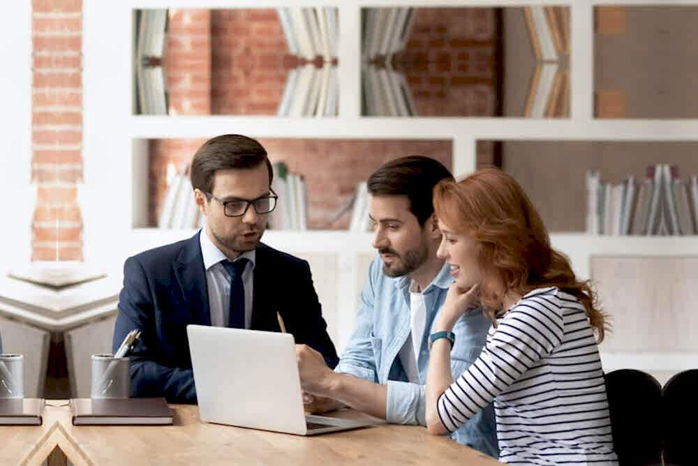 Three professionals are seated at a table in a modern office setting, with their faces obscured for privacy. One individual, dressed in business attire, is operating a laptop while the other two, also professionally dressed, are engaged in discussion with them. The environment suggests a collaborative work meeting focused on financial reporting.