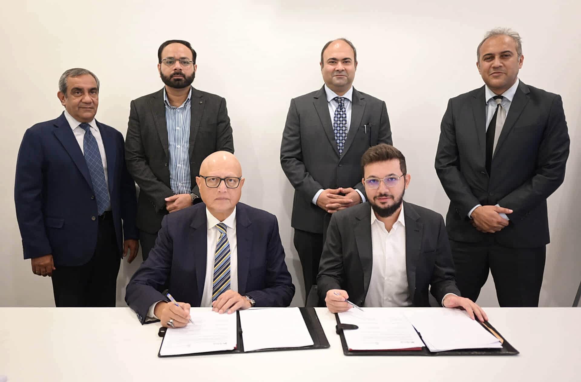 A group of six individuals, with their faces obscured for privacy, are present in a formal setting for a signing ceremony. Two individuals are seated at a table in the foreground, each signing a document, symbolizing the commencement of a partnership. Four individuals stand behind them, representing support and unity between the two organizations involved. The setting appears professional and is indicative of an important corporate event.