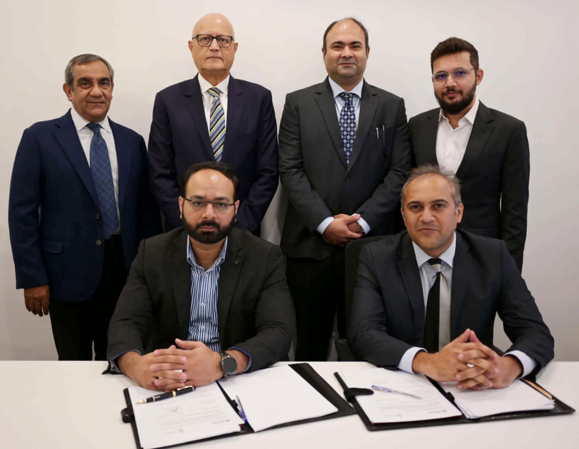 A signing ceremony with two seated individuals at the forefront signing documents and four standing individuals behind them in a professional office setting.