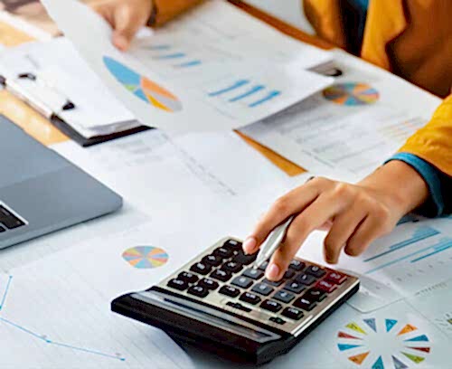 In a professional setting, a person’s hands are engaged in financial activities. One hand holds a document, while the other operates a calculator. The table displays various financial documents, including charts and graphs, suggesting work related to advisory and accounting services