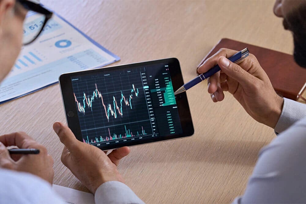 A person in a business setting is holding a stylus and interacting with a tablet displaying financial charts and graphs. The tablet screen shows a prominent upward-trending line graph among other analytical financial data. Papers with additional charts and data are visible on the desk, suggesting an environment of financial analysis or equity management.