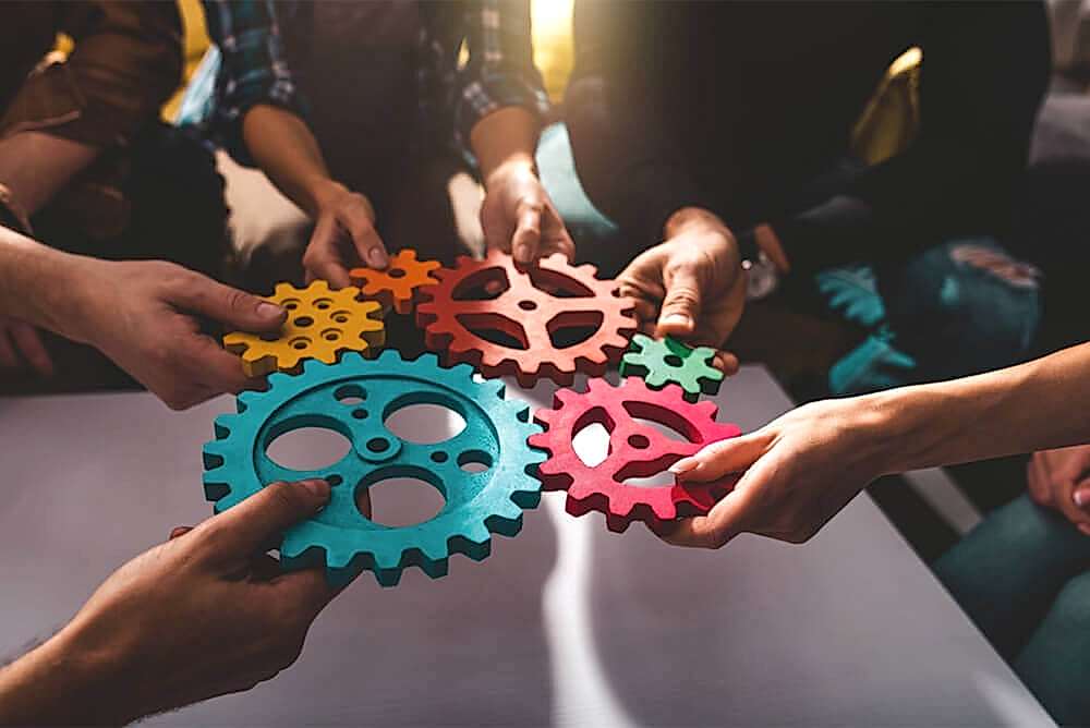 A group of hands coming together, each holding a colorful gear, symbolizing teamwork and integration in a business setting. The gears are interlocking, suggesting the seamless merging of companies or systems as referenced by IFRS 3 Business Combination.