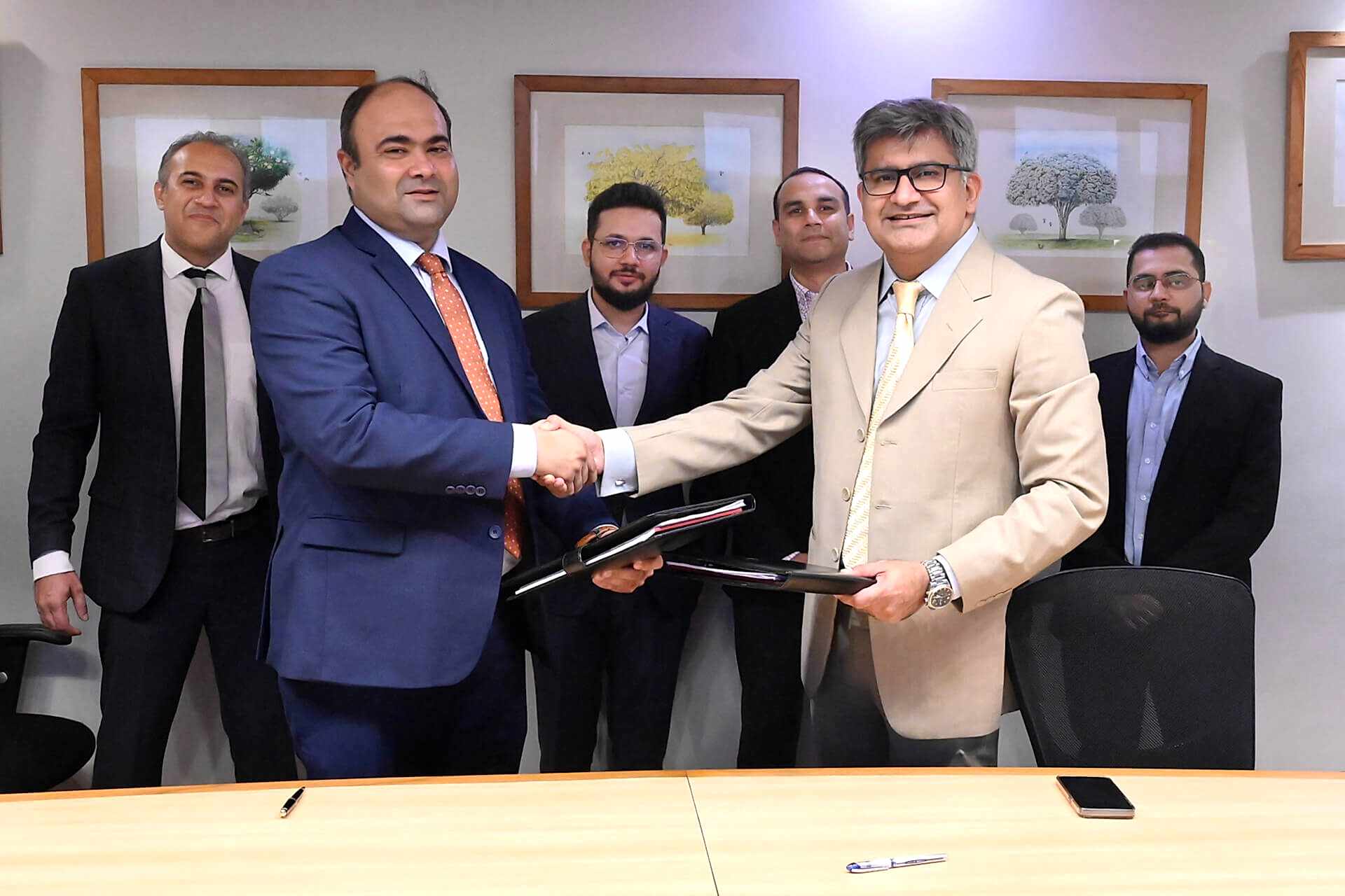 Two professionals, identified as Faisal Khan (CEO of IGI Insurance) and Ibrahim Zahidie (Director Advisory & Transformation at Prima Consulting), are shaking hands in the foreground of a conference room. Behind them stand four individuals: Shabih Ahmed Arif (Director Actuarial Services), Sumar Sayani (Director AI & Technology), Syed Awais Amjad (CFO), and Murtaza Feroz Jandga (Finance Controller) from IGI Insurance and Prima Consulting. They are all facing the camera with obscured faces for privacy. The setting suggests a formal business agreement or partnership.