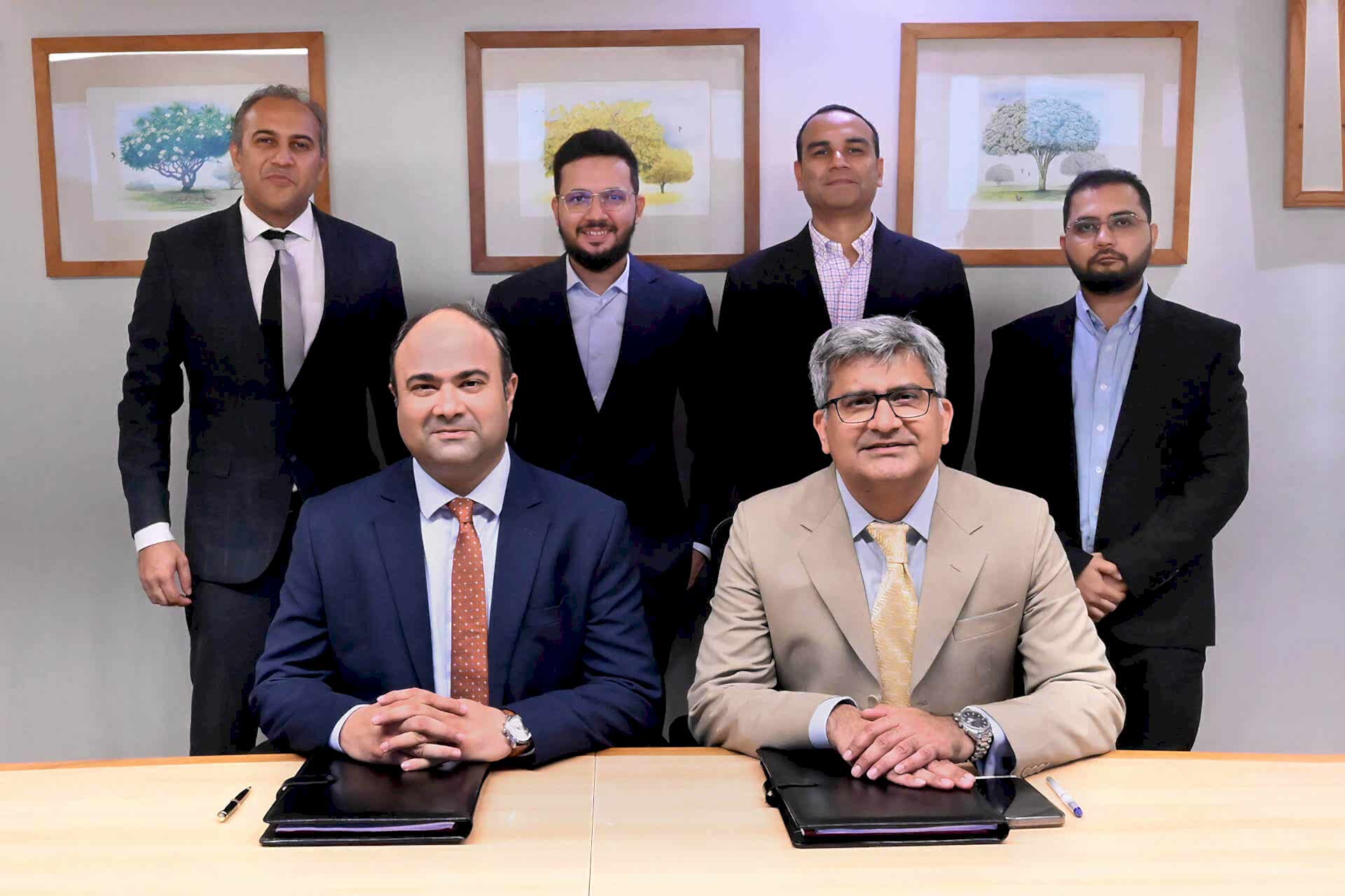 The image captures a formal business setting where two individuals, identified as Faisal Khan (CEO of IGI Insurance) and Ibrahim Zahidie (Director Advisory & Transformation at Prima Consulting), are seated at the forefront with documents on the table, symbolizing a partnership agreement. Behind them stand four professionals: Shabih Ahmed Arif (Director Actuarial Services at Prima Consulting), Sumar Sayani (Director AI & Technology at Prima Consulting), Syed Awais Amjad (CFO of IGI Insurance), and Murtaza Feroz Jandga (Finance Controller at IGI Insurance). All individuals are professionally dressed, and the setting suggests a significant corporate event.