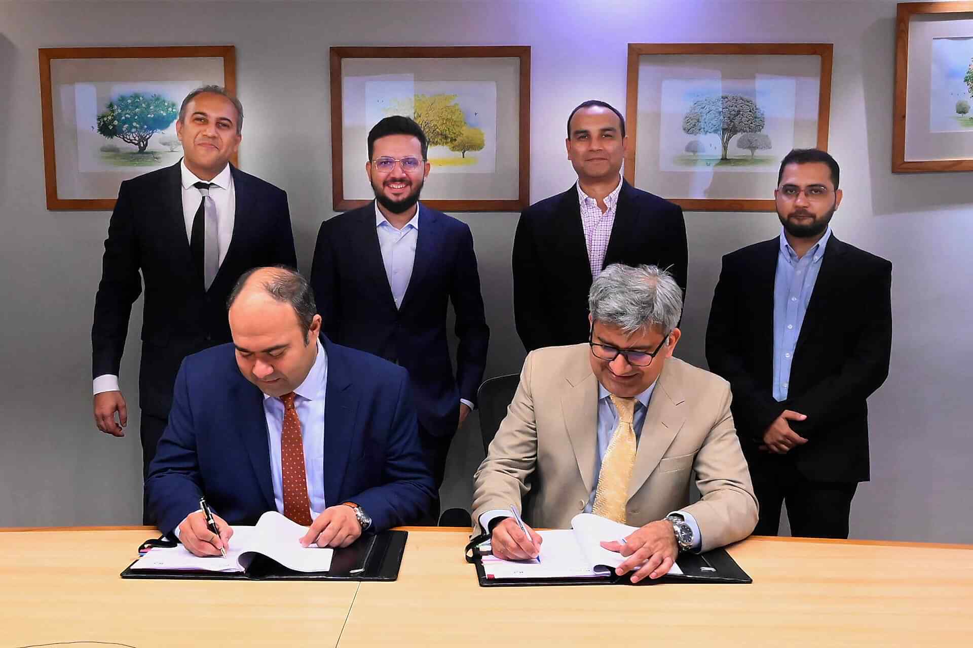 The image captures a significant moment where Faisal Khan, CEO of IGI Insurance, and Ibrahim Zahidie, Director Advisory & Transformation at Prima Consulting, are seated at a table signing documents to formalize their partnership. Behind them stand Shabih Ahmed Arif, Director Actuarial Services at Prima Consulting; Sumar Sayani, Director AI & Technology at Prima Consulting; Syed Awais Amjad, CFO of IGI Insurance; and Murtaza Feroz Jandga, Finance Controller at IGI Insurance. The individuals’ faces are not visible. The setting suggests a professional environment with artwork on the wall, indicating the importance of the event.