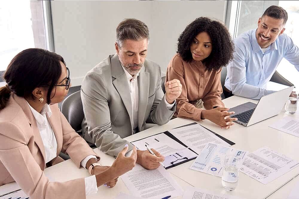 A professional meeting in progress featuring four individuals around a table, with their faces obscured for privacy. One person is taking notes, another is gesturing towards a document, and the others are attentively engaged in the discussion. Visible on the table are papers with graphs and charts, a laptop, and a bottle of water, indicating an analytical or strategic business environment. This image likely represents the collaborative and expert nature of Prima Consulting’s Financial & Risk Advisory Services