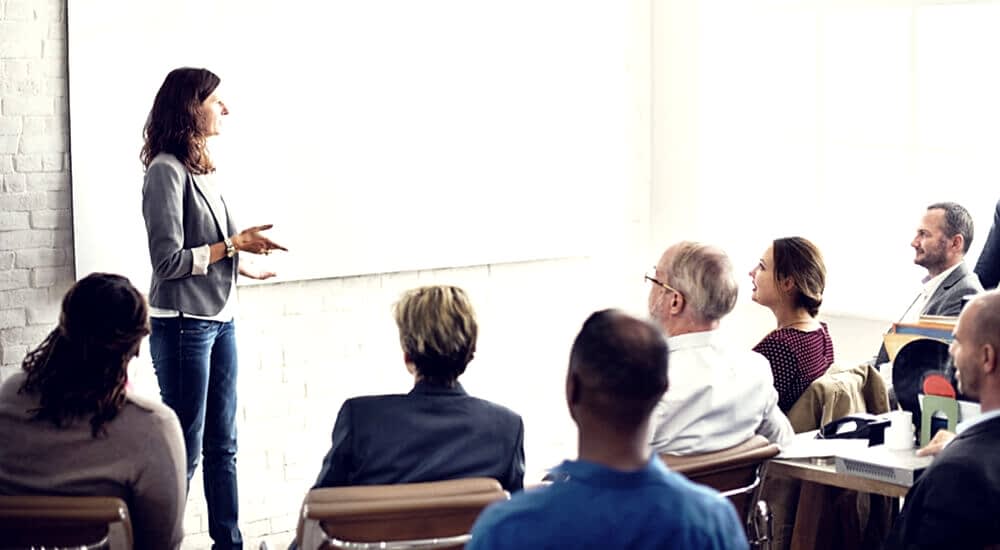 A professional training session in a bright room with large windows. An instructor, facing away from the camera, is gesturing towards a blank projection screen while addressing attendees seated in rows. The attendees are focused on the instructor, suggesting an engaging educational environment.