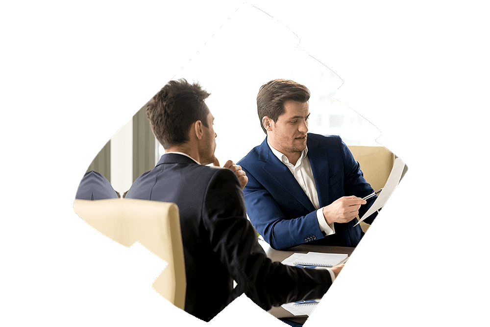 Two individuals with obscured faces are engaged in a professional review session. One individual is seated at a table with an open laptop, facing the other who is holding and examining a document. The setting suggests a formal business environment, likely discussing financial or audit-related matters.