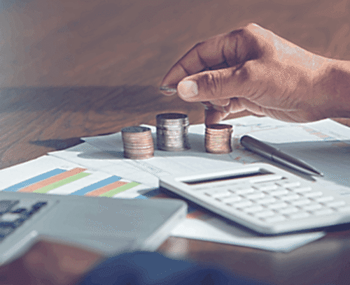 A close-up image of a hand stacking coins in ascending order on a desk with financial charts, a calculator, and additional coins laid out. The scene suggests financial planning or analysis.