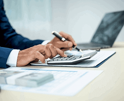 A close-up image of a person’s hands using a calculator and holding a pen, with documents and a laptop in the background, suggesting financial work or analysis.