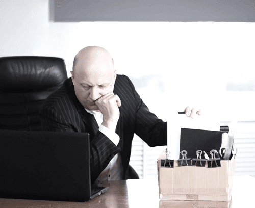 An individual in a business suit is seated at a desk, facing away from the camera, with their face obscured for privacy. The person appears to be in an office setting, holding a document or folder while gesturing with the other hand as if in discussion or explanation. A briefcase and what seems to be a laptop are also visible on the desk, suggesting a professional environment.