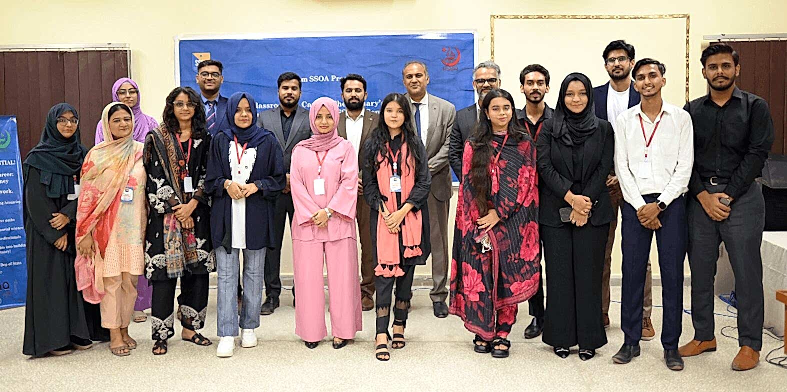 A group of individuals stands together in a room, posing for a group photo. They are dressed in a variety of professional and semi-formal attire, with some wearing traditional clothing. The background features a blue banner and informational posters, indicating an event or gathering.